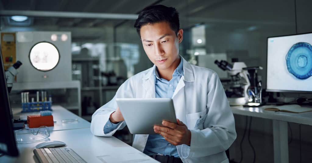 A scientist in a lab coat sits at a desk, looking at a tablet. A microscope and computer monitor are visible in the background.