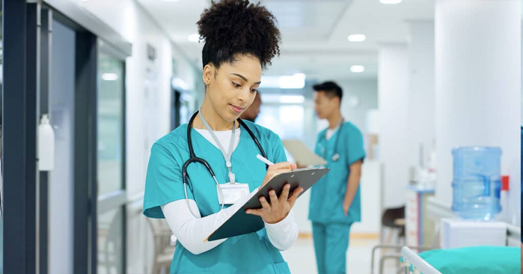 A healthcare professional in scrubs writes on a clipboard in a hospital corridor, with another person in scrubs in the background.