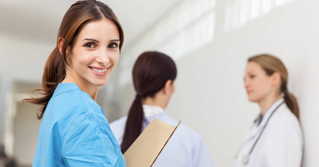 A woman in a blue scrubs top smiles while holding a folder, with two colleagues in white coats talking in the background.