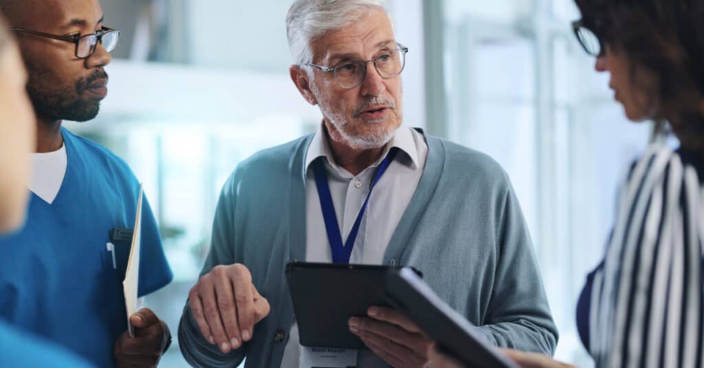 An elderly man with glasses speaks to three people, including a doctor. He holds a tablet, with a focused expression. The setting is indoors, possibly a medical or professional environment.