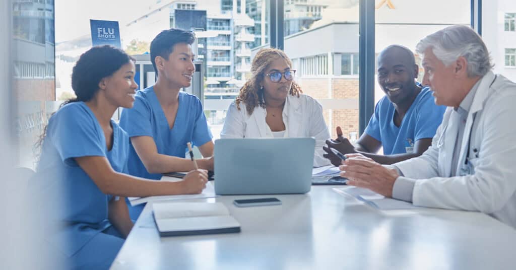 Doctors and medical staff, showcasing their medical expertise, engage in discussion around a table with laptops and notebooks in a bright room. A "Flu Shots" sign is visible in the background.
