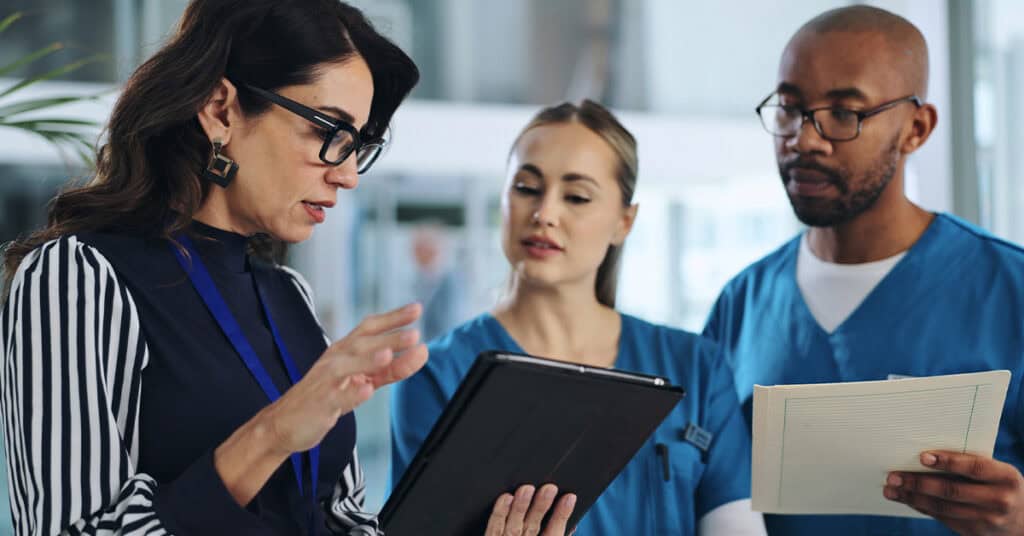 A woman holding a tablet talks to two people in blue scrubs.