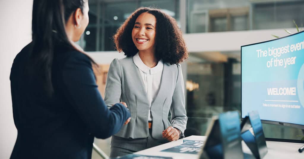 Two professionals shake hands at a reception desk with promotional materials and a display screen in the background.