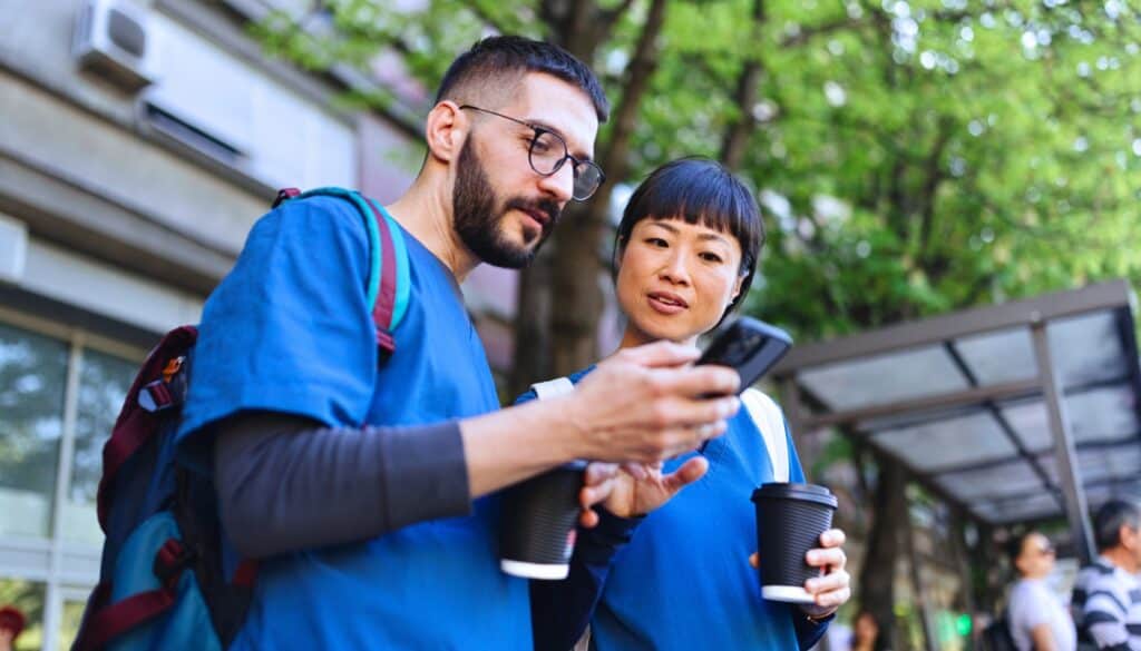 Two healthcare professionals in blue scrubs, possibly travel nurses, hold coffee cups while looking at a smartphone outside a building with greenery and people bustling in the background.