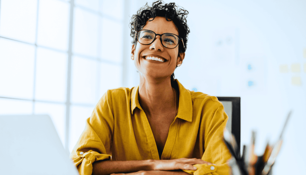 A person wearing glasses and a yellow shirt smiles while sitting at a desk with a laptop and pencils, near a window, reflecting on their journey as a traveling social worker.