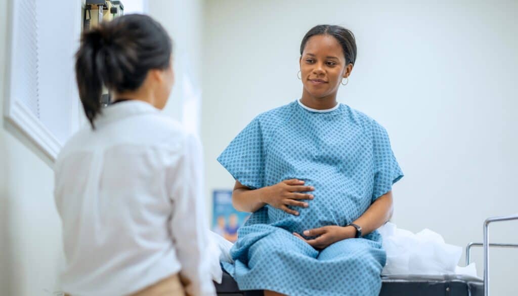 A pregnant woman in a hospital gown sits on an examination table, holding her belly. She is speaking to a healthcare professional.