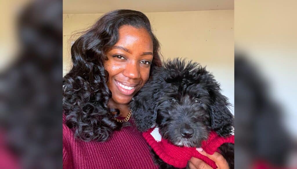 A woman with long, wavy hair smiles and poses indoors with a black, curly-haired puppy wearing a red sweater.