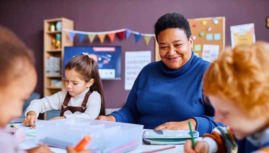 A teacher sits with children at a table in a classroom. The children focus on drawing with colored pencils. A bulletin board and colorful bunting are in the background.