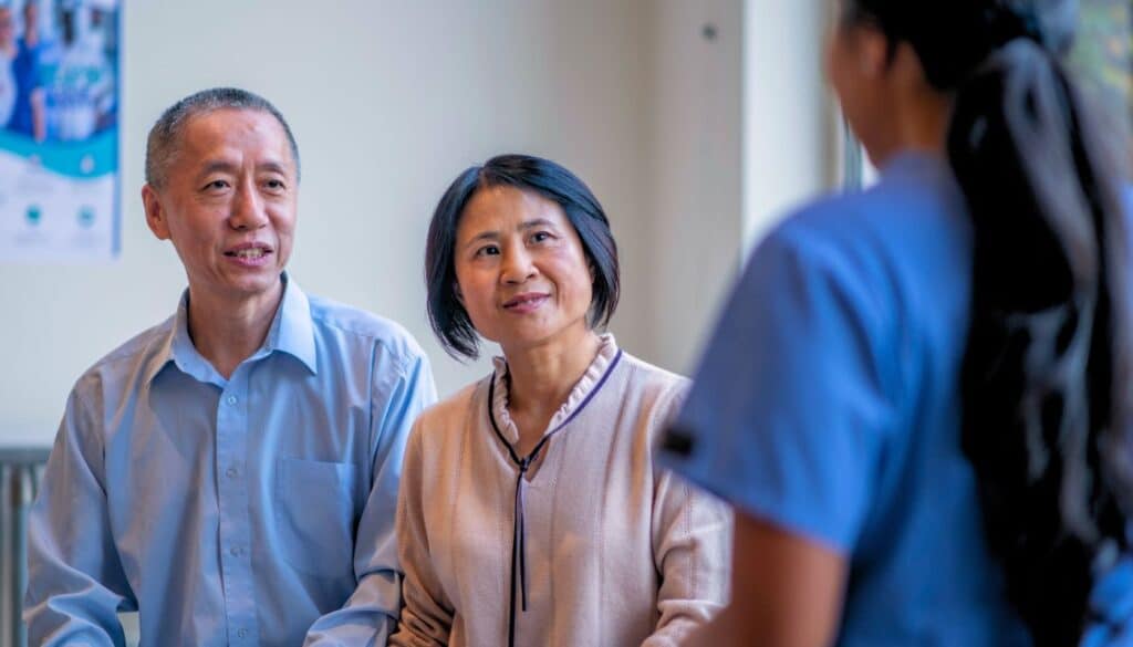 A man and woman sit together, attentively listening to a healthcare professional in blue scrubs.