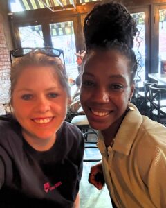 Two women smiling indoors at a restaurant or café, seated closely together. Natural light comes through large windows behind them.