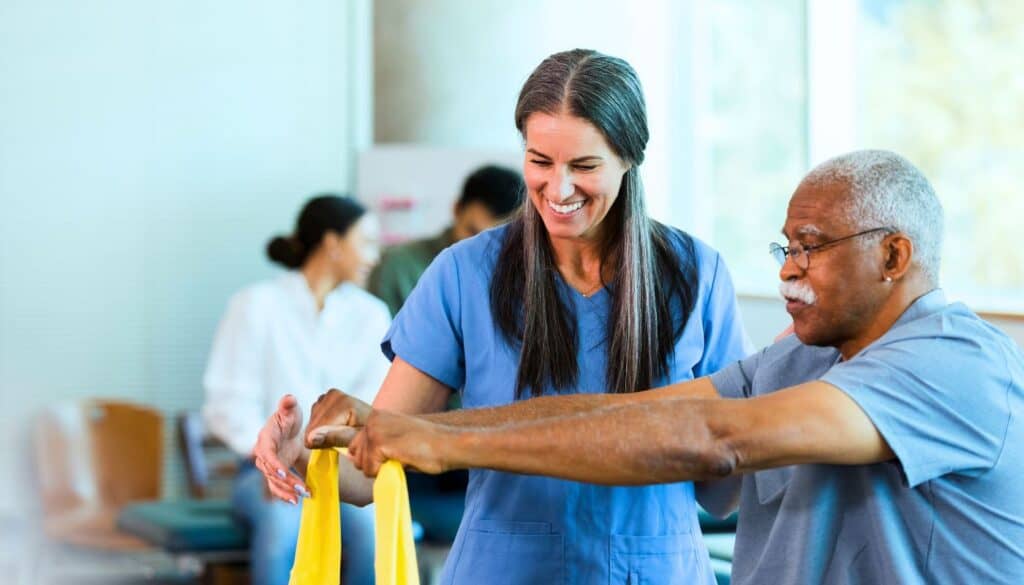 A physical therapist assists an older man with arm exercises using a yellow resistance band in a medical rehabilitation setting.