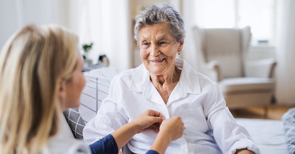 A young woman nurse assists an older woman by buttoning her shirt while they sit together in a bright living room.
