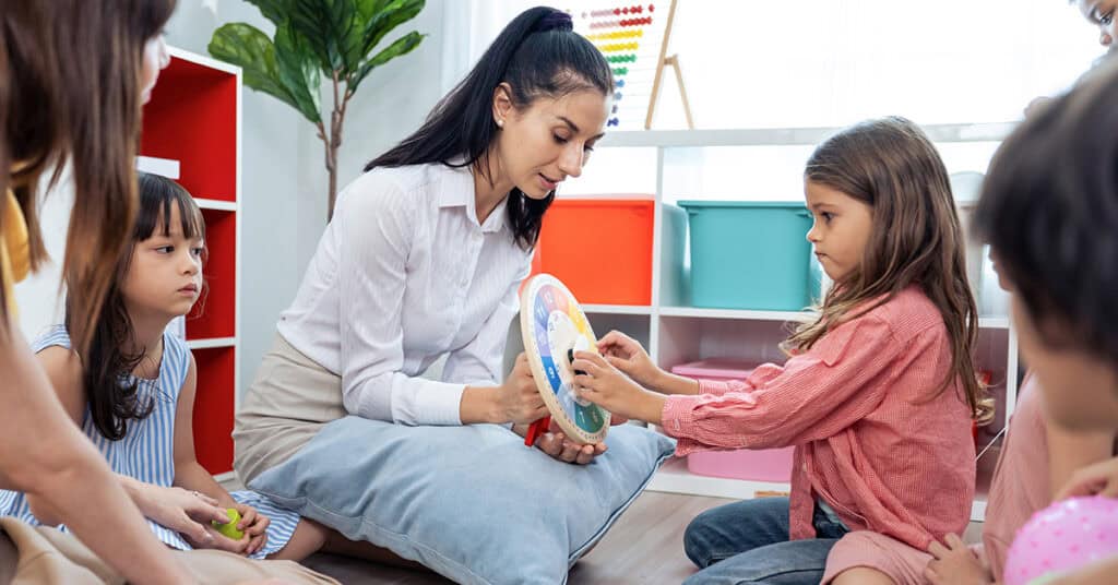 A woman sits on the floor with children, holding a colorful learning clock while a girl in a pink shirt interacts with it. Other children watch attentively in a classroom setting.