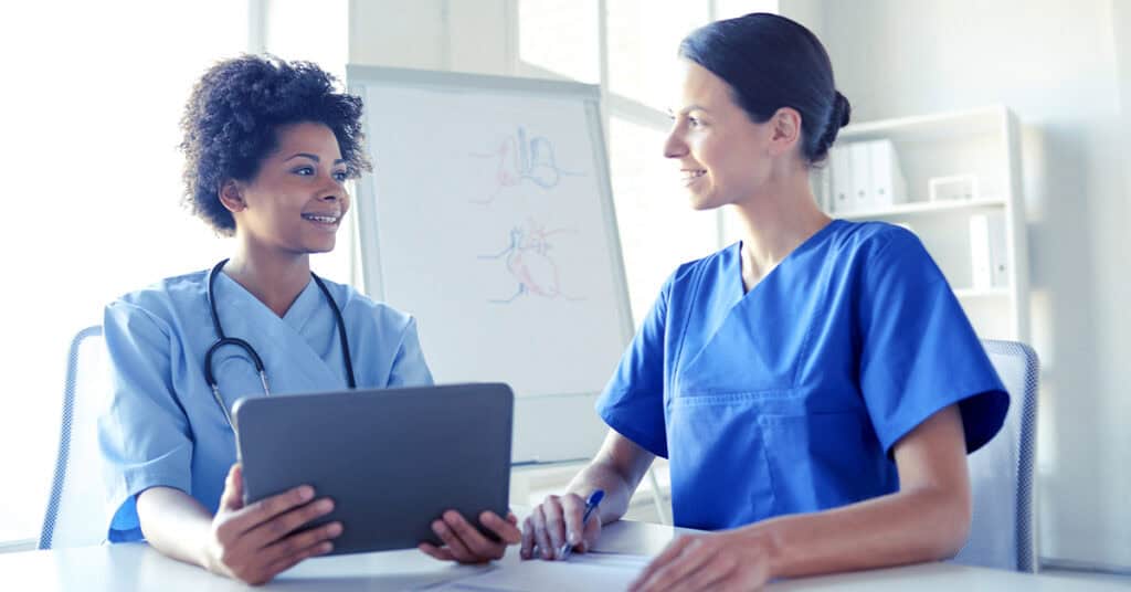 Two healthcare professionals in scrubs discuss information, one holding a tablet and the other taking notes, in front of a whiteboard with anatomical diagrams.