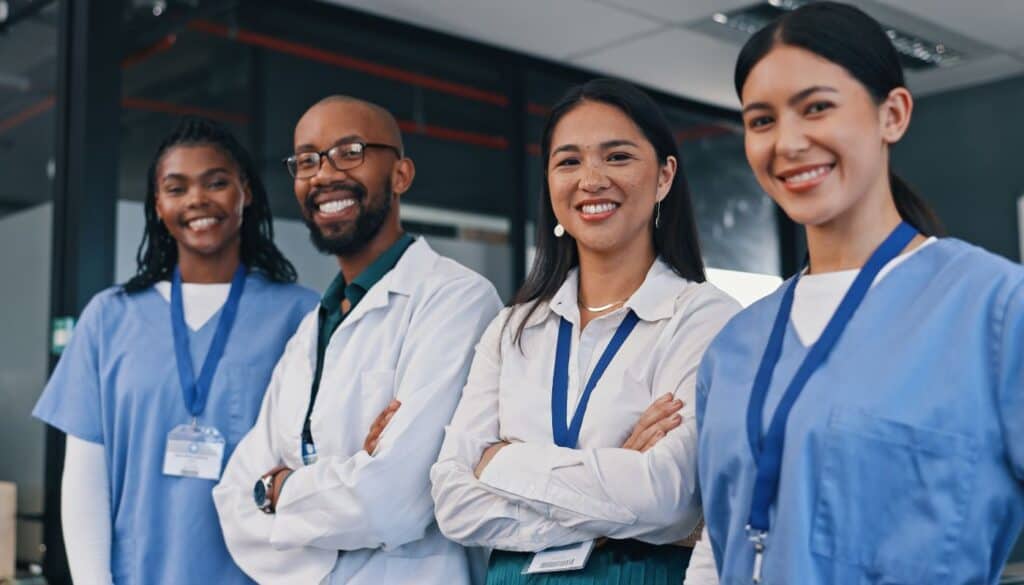 Four healthcare professionals, two in white coats and two in blue scrubs, stand together indoors, smiling at the camera.