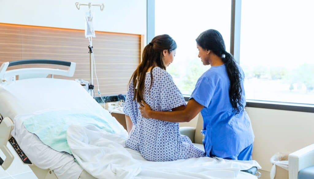 A nurse assists a patient in a hospital gown as she sits on the edge of a hospital bed in a bright, modern room.