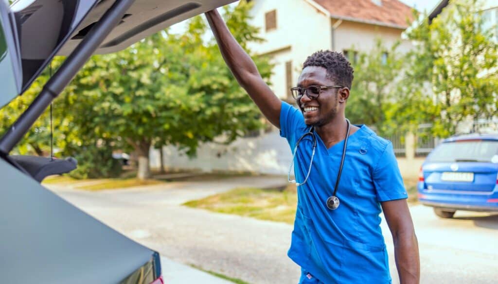A person in blue medical scrubs with a stethoscope smiles while loading or unloading items from a car trunk in a residential area.
