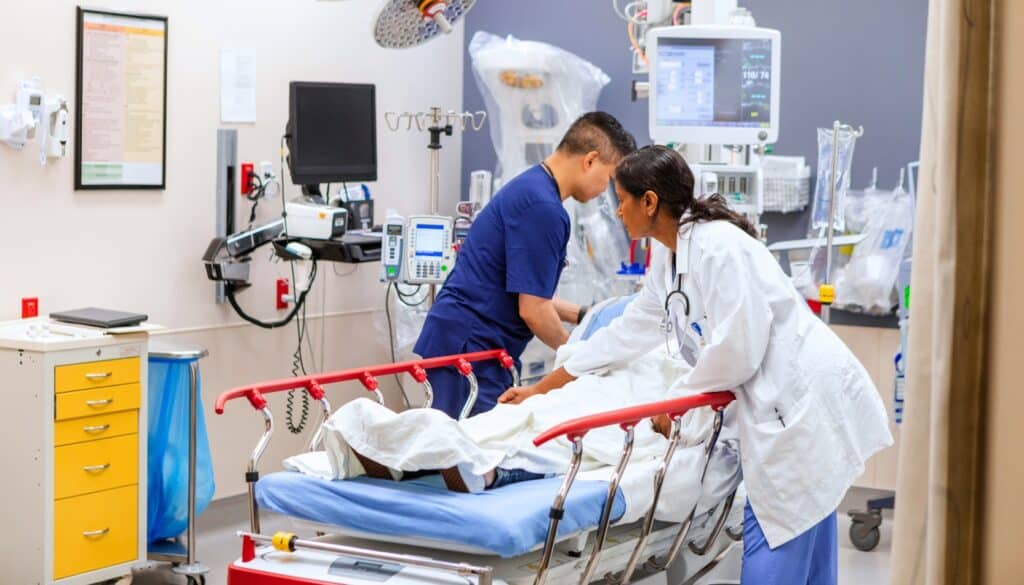 Two healthcare professionals attend to a patient lying on a hospital bed in a medical examination room with equipment and monitors.