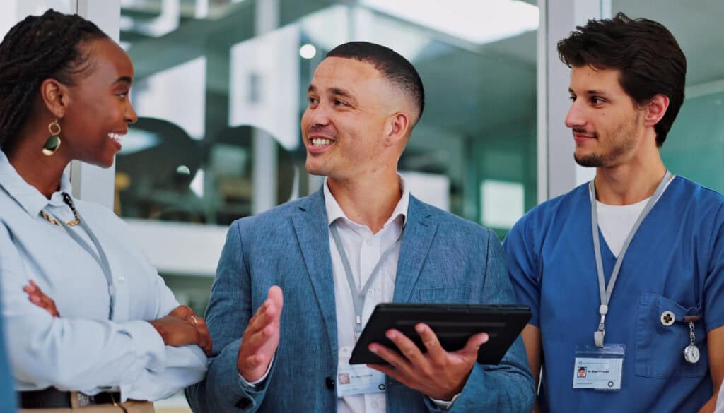 Three healthcare professionals converse indoors; one holds a tablet and gestures while the others listen attentively.