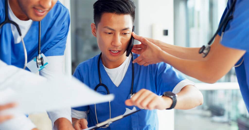 A healthcare worker in blue scrubs checks his watch while talking on the phone, with colleagues and medical documents nearby.