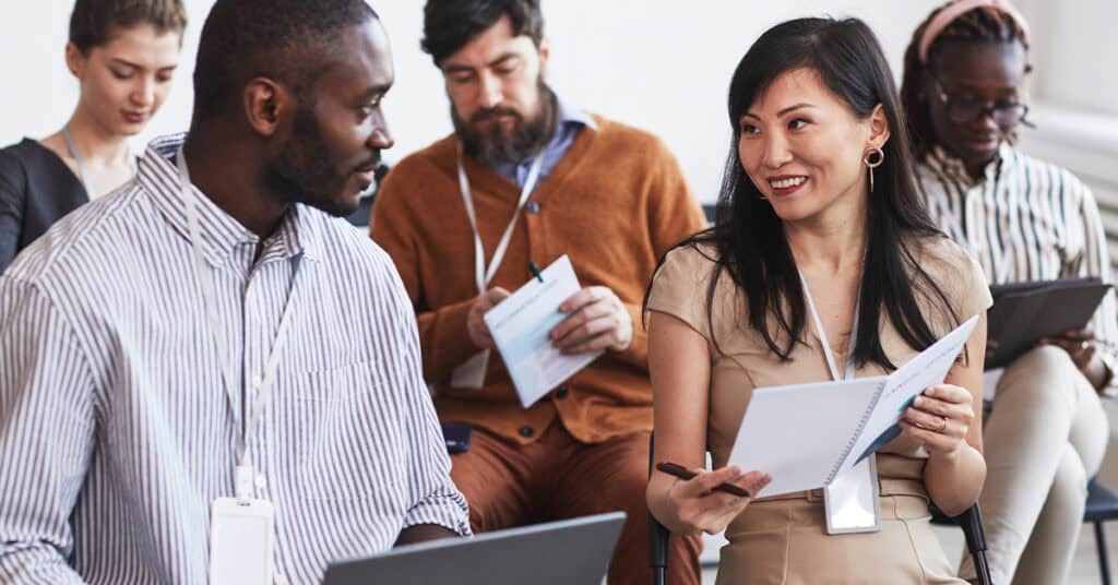 A group of adults sits in a meeting room. Two people in front are talking and holding documents, while others listen, take notes, or look at electronic devices.