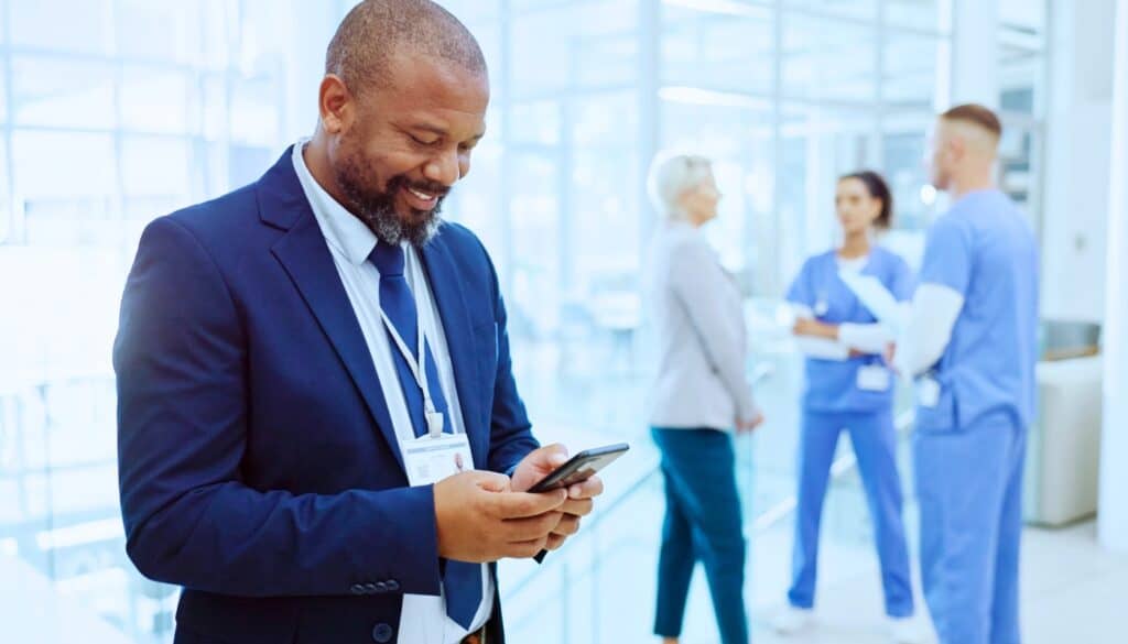 A man in a suit with a hospital badge looks at his phone and smiles, while three medical staff talk in the background of a bright, modern hospital setting.