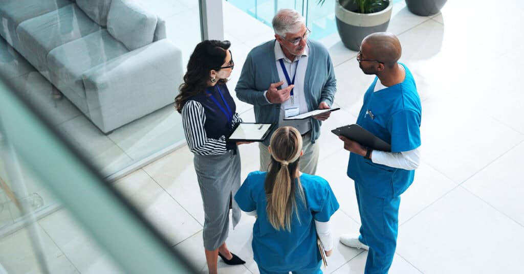 Four healthcare professionals, two in scrubs and two in business attire, stand in a hospital corridor having a discussion while holding clipboards and a tablet.