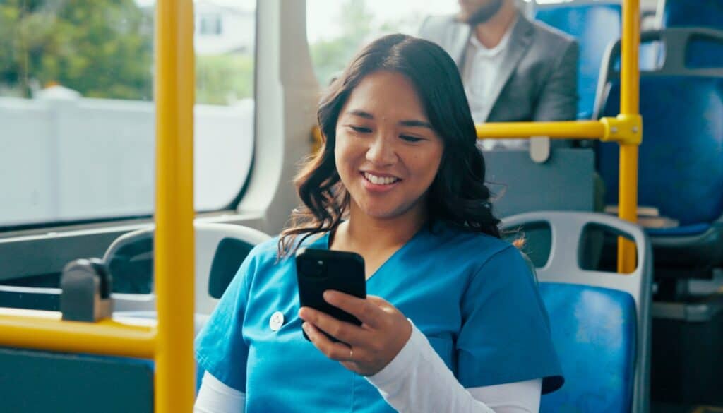A woman in blue scrubs sits on a bus, smiling while looking at her smartphone.