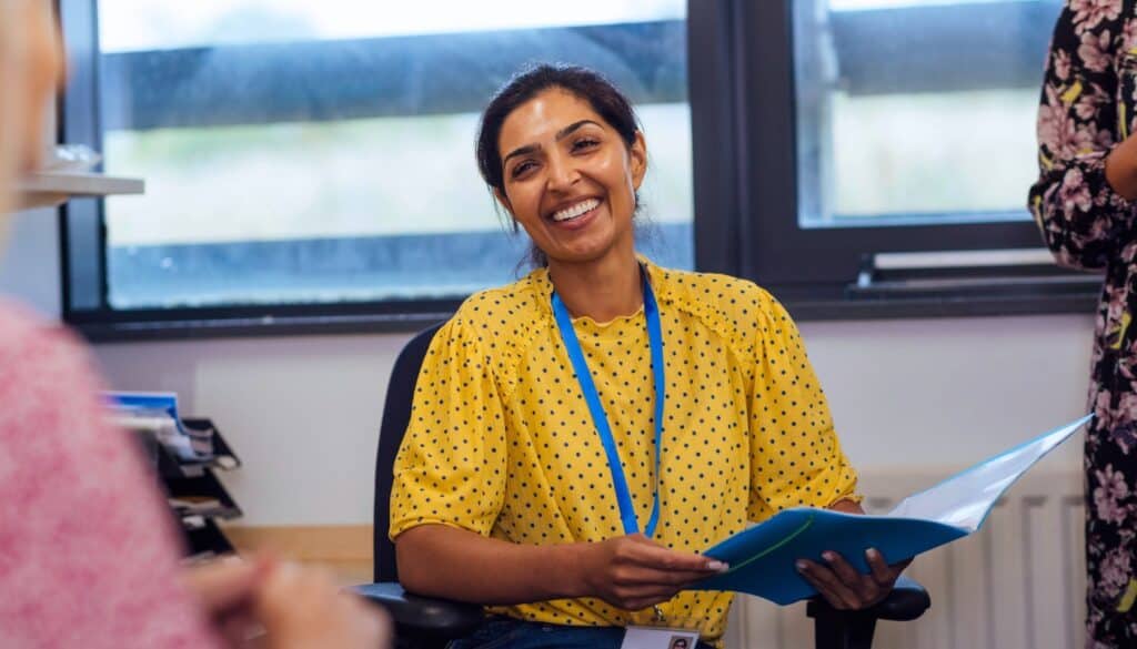 A woman in a yellow polka dot blouse sits in an office chair, smiling and holding an open folder, with a lanyard around her neck.