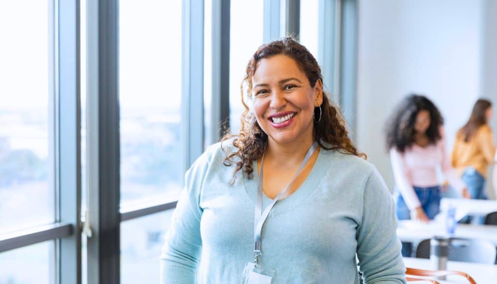 A woman with curly hair, wearing a light blue sweater and lanyard, smiles while standing in a bright office with large windows; people are in the background.