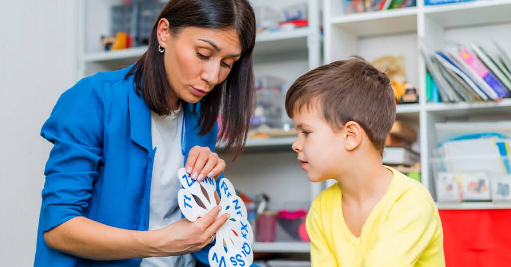 A woman shows a round card with letters to a young boy in a classroom or therapy setting, with shelves of educational materials in the background.