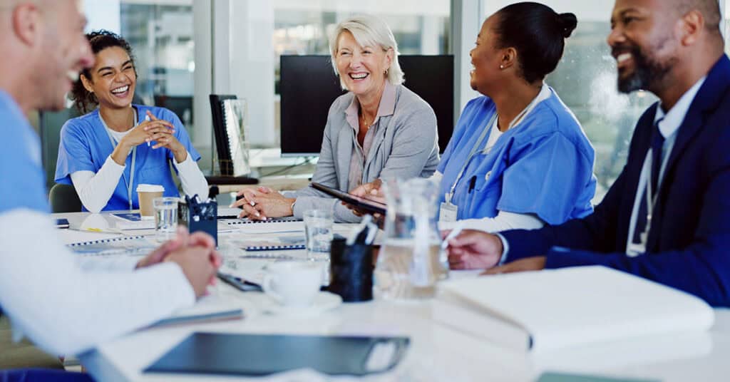 A group of healthcare professionals and an administrator sit around a conference table, engaged in a discussion and smiling.