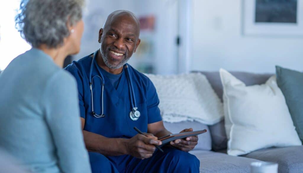A healthcare professional in blue scrubs with a stethoscope talks to a seated patient on a couch, holding a tablet and smiling.