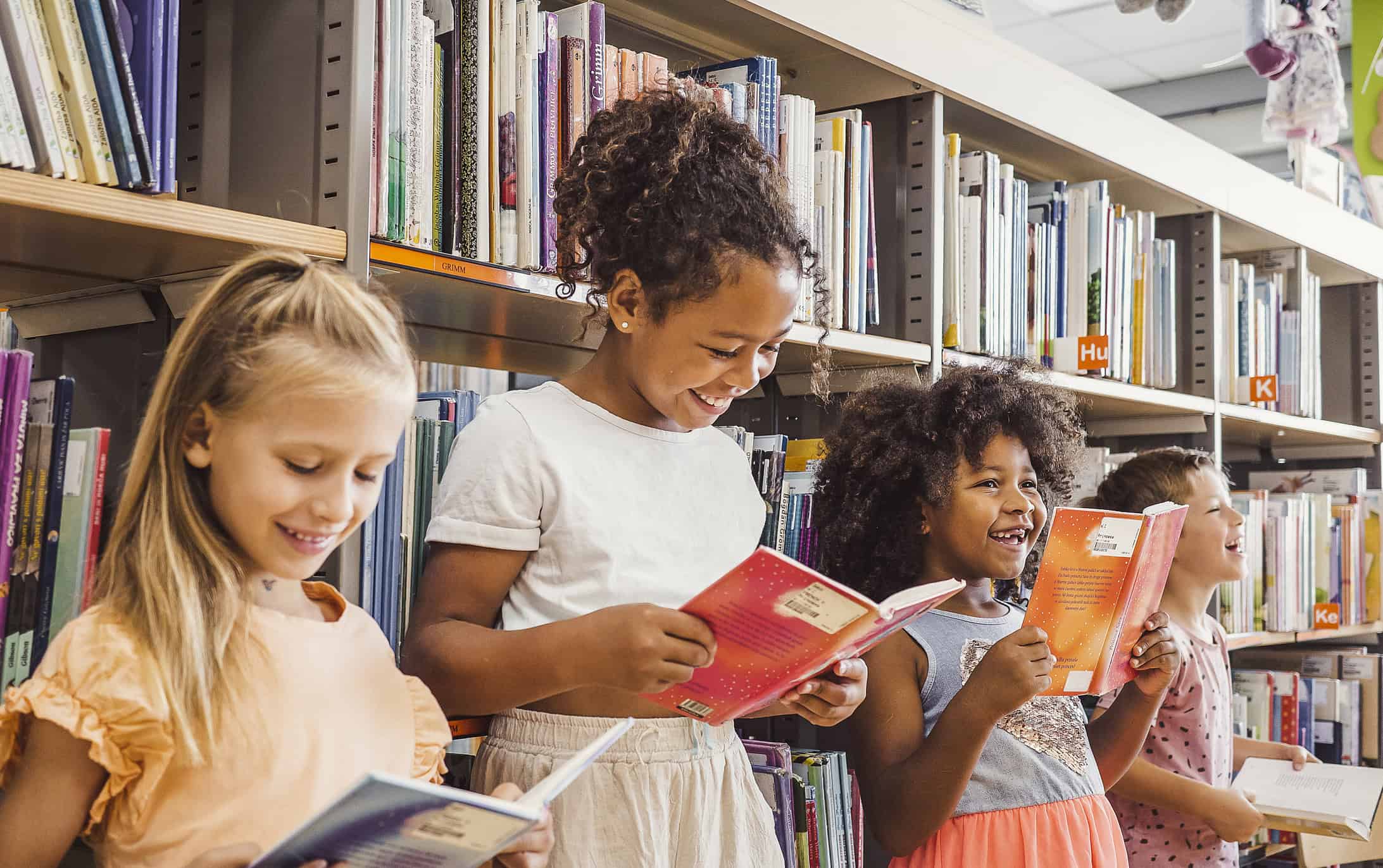Four children stand in front of bookshelves in a library, smiling and reading books.