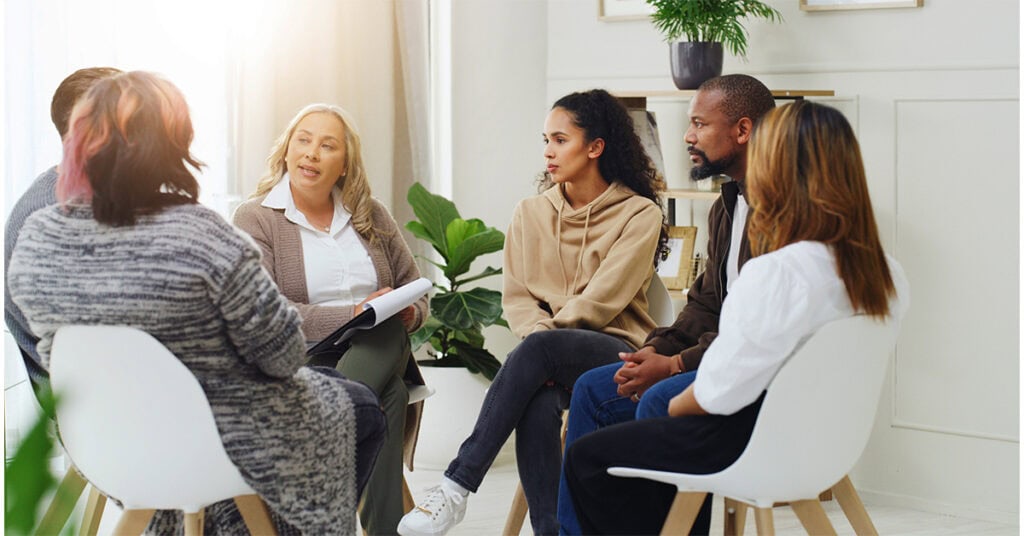 A group of five people sit in a circle having a discussion in a well-lit room with plants and framed pictures.