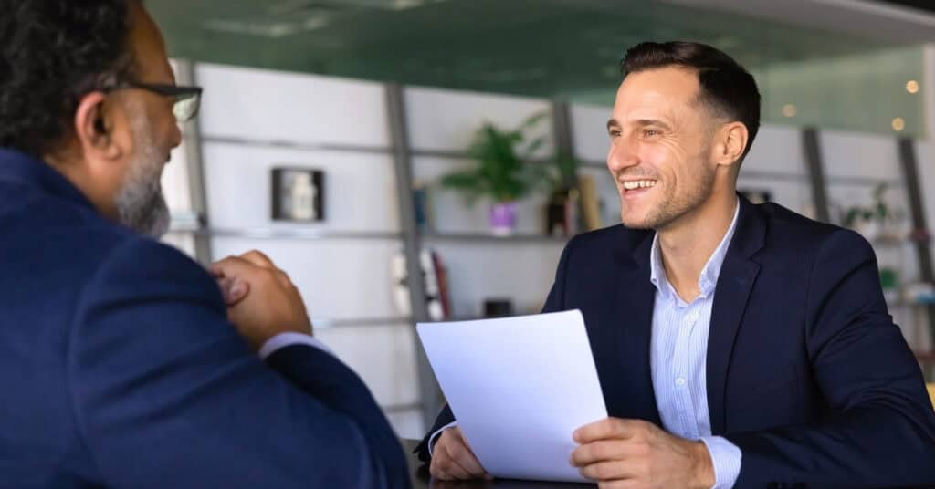 Two men in business attire sit across from each other at a table, one holding a sheet of paper and smiling during a professional meeting or interview.