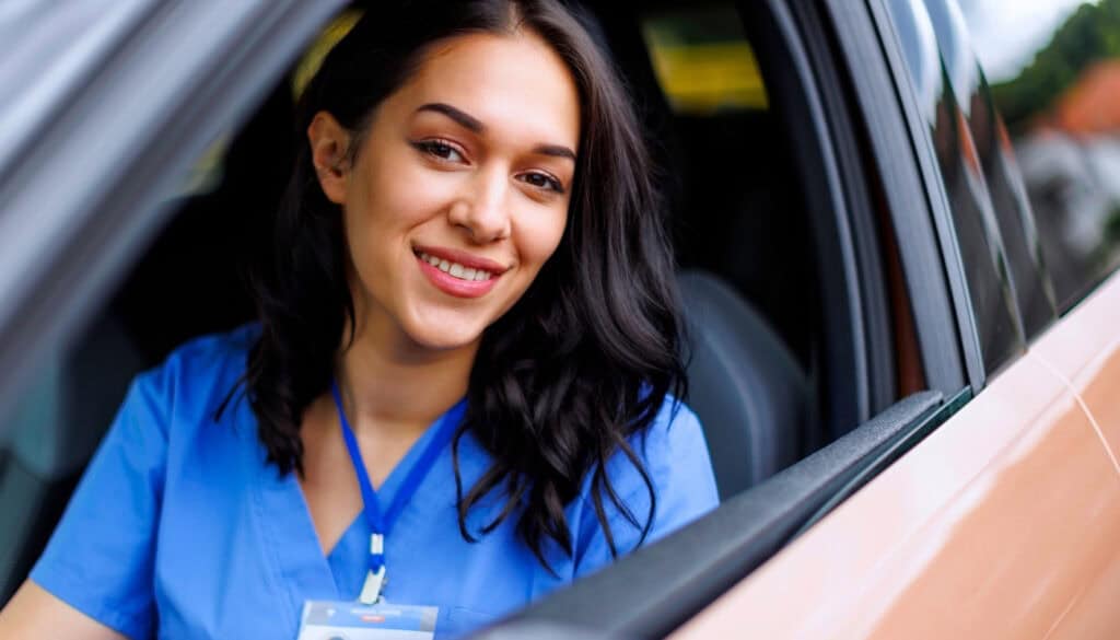 A woman wearing blue scrubs and a name badge sits in a car, smiling at the camera.