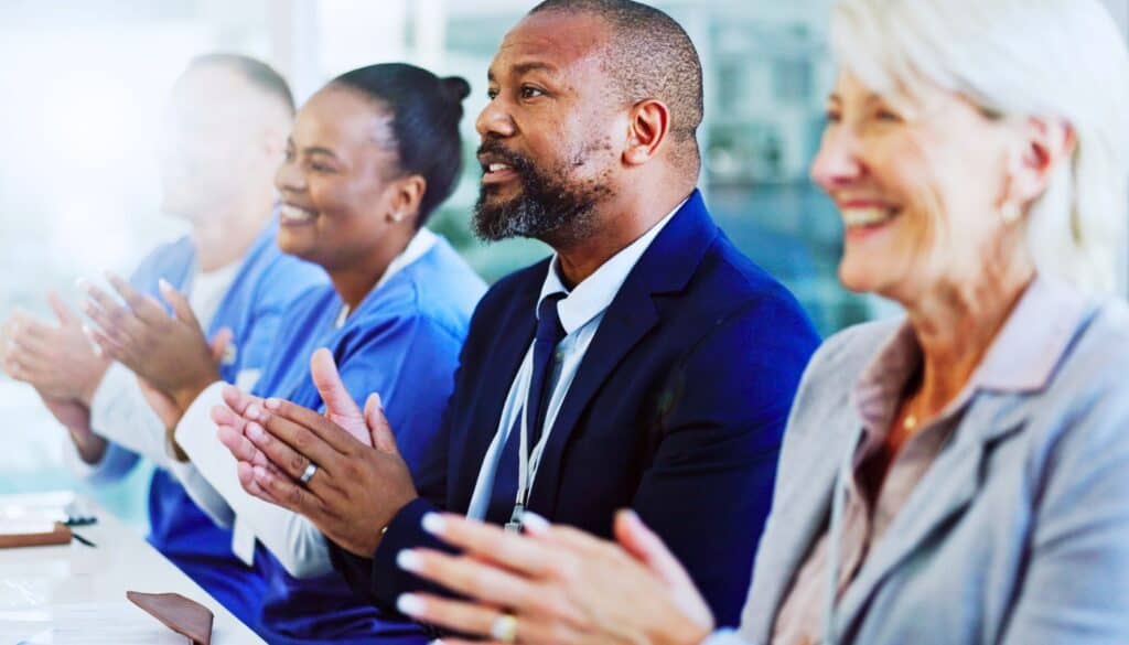 Four professionals, including medical staff and business personnel, sit in a row and applaud during a meeting or presentation in a bright, modern setting.
