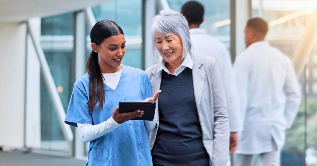 A nurse in scrubs shows a tablet to an older woman in business attire in a modern medical facility, with other medical staff visible in the background.