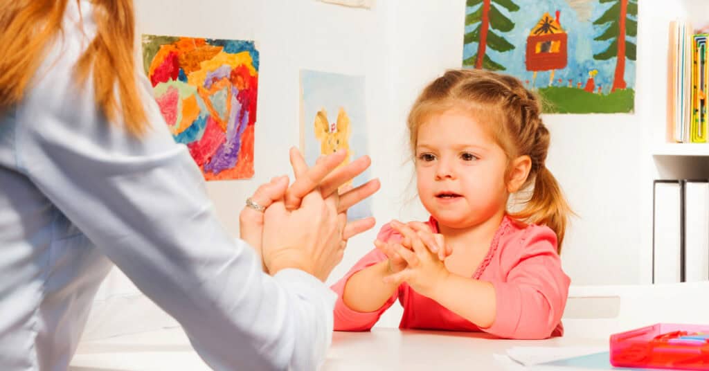 A young girl and an adult sit at a table, engaging in a hand exercise. Children's artwork is visible on the wall behind them.
