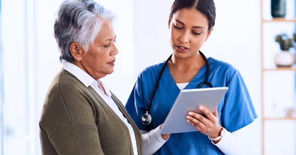 A healthcare professional in scrubs shows a tablet to an older woman, discussing information in a medical setting.