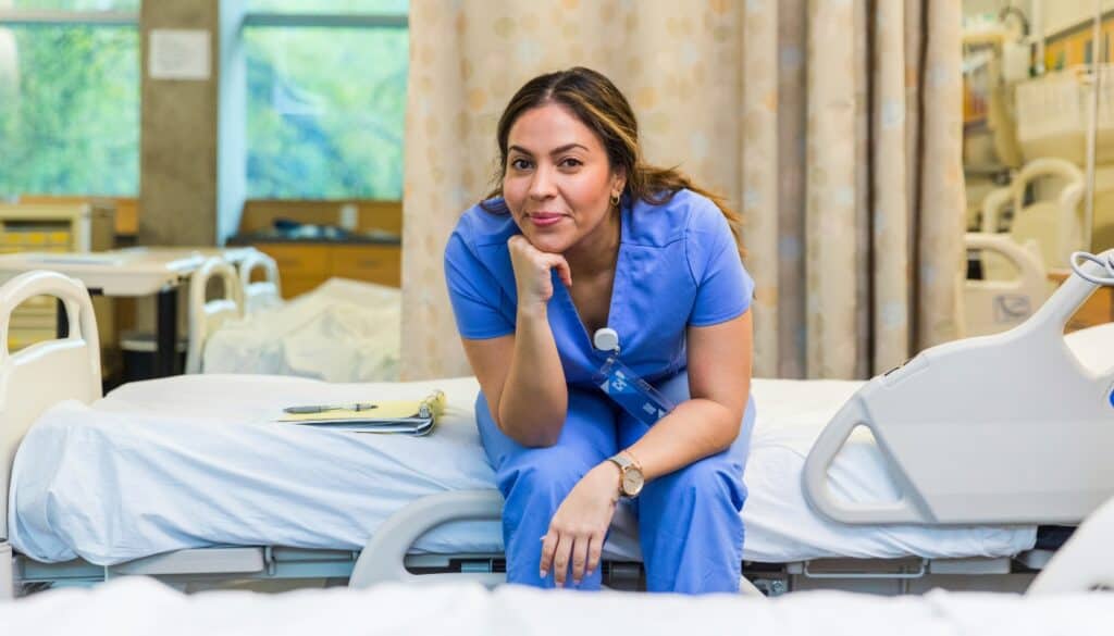 A nurse in blue scrubs sits on the edge of a hospital bed, resting her chin on her hand, with a clipboard nearby in a well-lit hospital room.