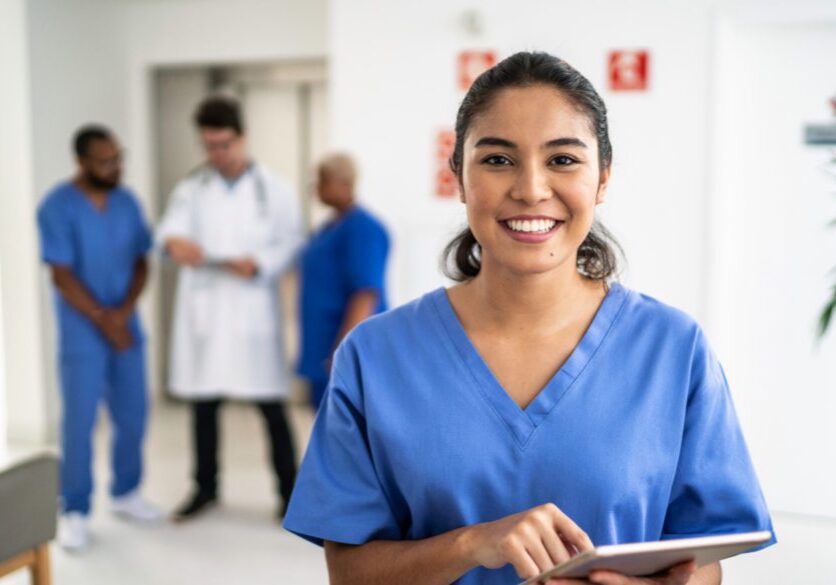 photo of a nurse smiling at the camera with three healthcare professionals talking in the blurred background